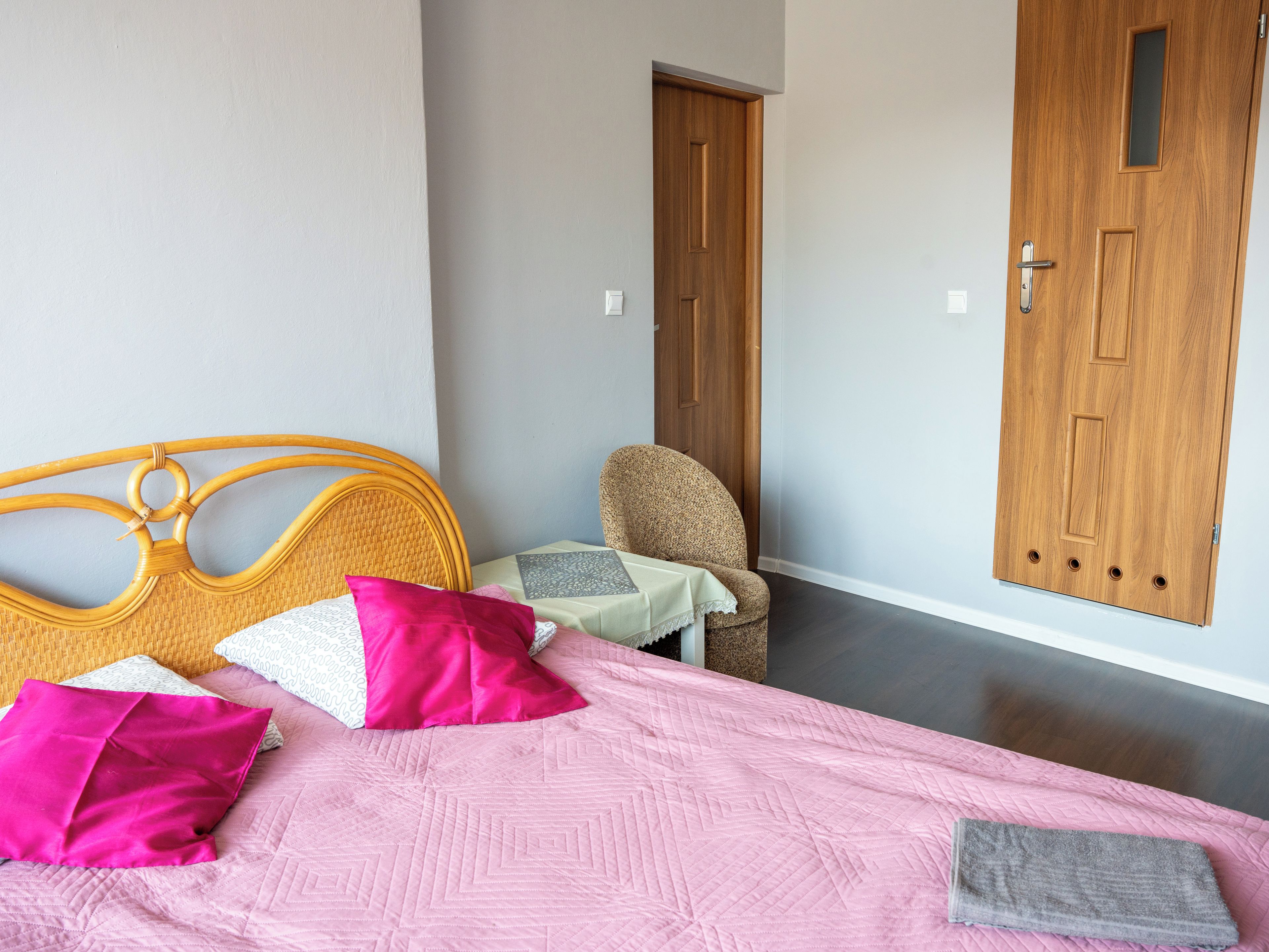 A bedroom with a bed, pillows, a chair, a small table, and two wooden doors.