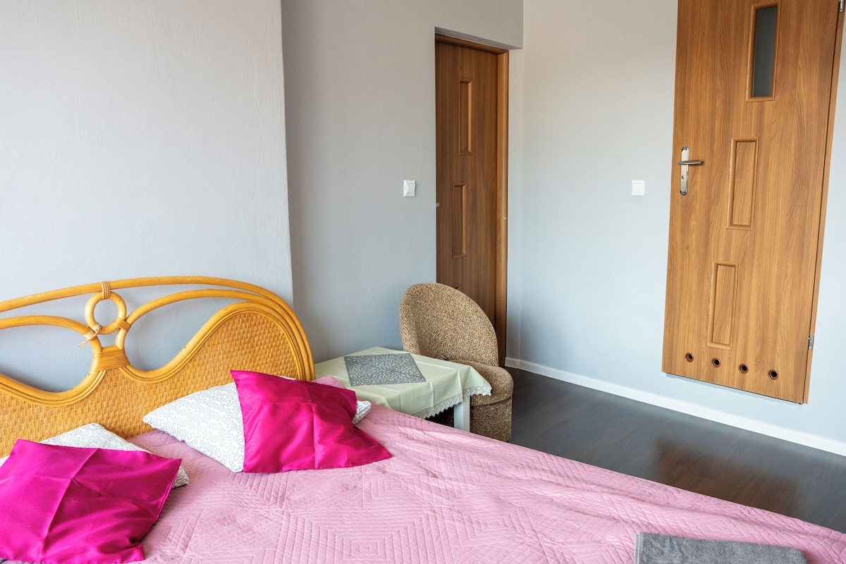 A bedroom with a bed, pillows, a chair, a small table, and two wooden doors.