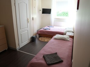 A bedroom with two beds, a TV mounted on the wall, a radiator, and a window with a view of greenery. - La Perla (Międzyzdroje)