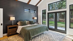A bedroom with a large bed, two bedside tables with lamps, a dark wall, and large windows with a view of greenery.