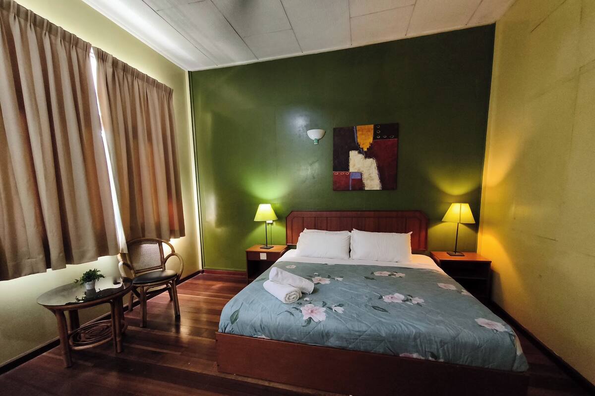 Ein Hotelzimmer mit einem Bett, zwei Nachttischlampen, einem Sessel, einem kleinen Tisch mit Pflanze und einem Bild an der Wand.