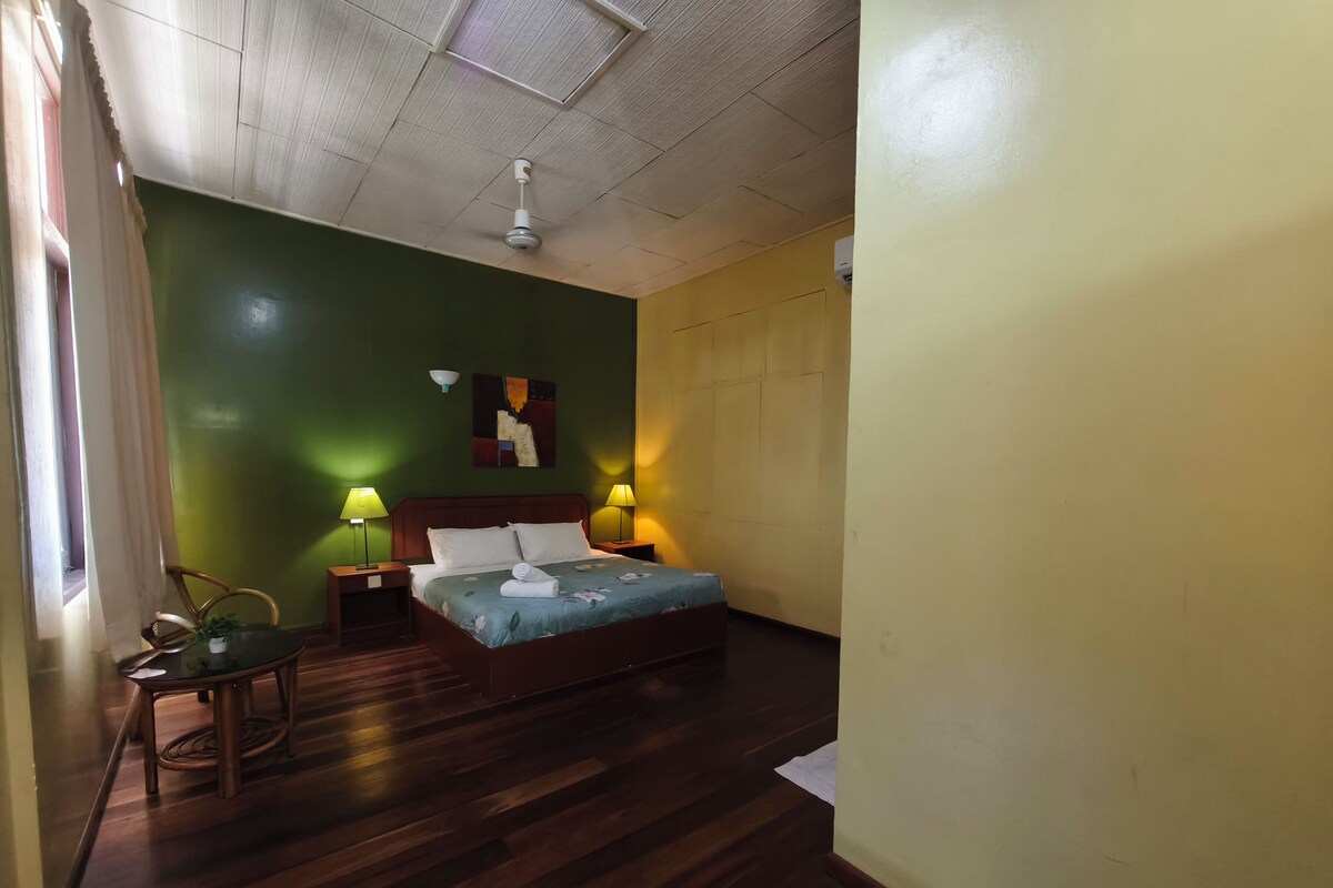A bedroom with a bed, two bedside lamps, a ceiling fan, a green wall, a wooden floor, and a window with curtains.