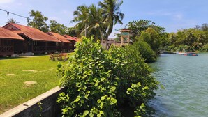A riverside view with a grassy area, a wooden building with a red roof, and a sign for 'Qurata Riverside Resort'. - Qurata Riverside Resort (Kuala Terengganu)