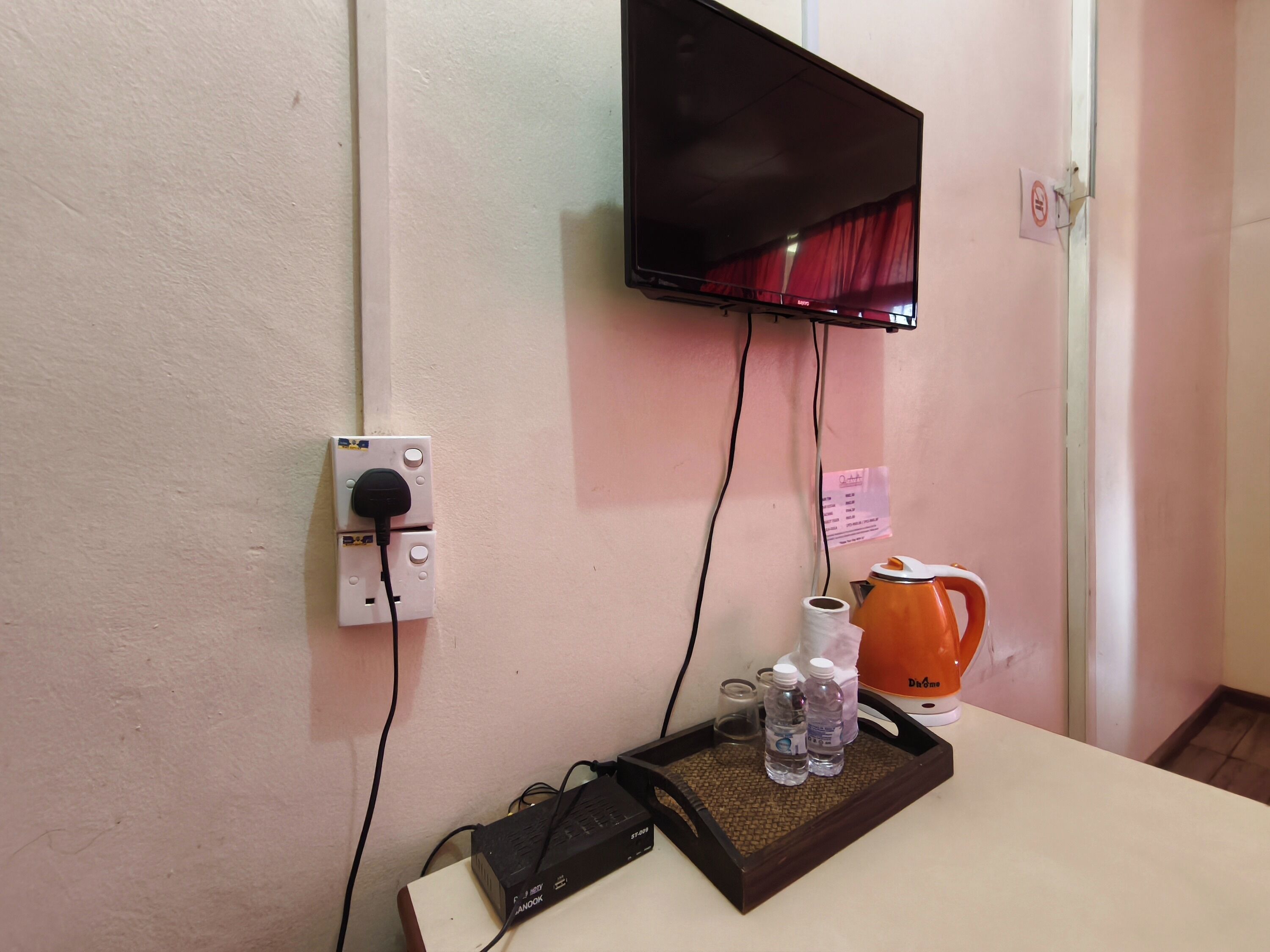 Une chambre avec un téléviseur fixé au mur, une prise de courant, un plateau avec des bouteilles et une bouilloire, et une porte sur laquelle est apposée une pancarte « accès interdit ».