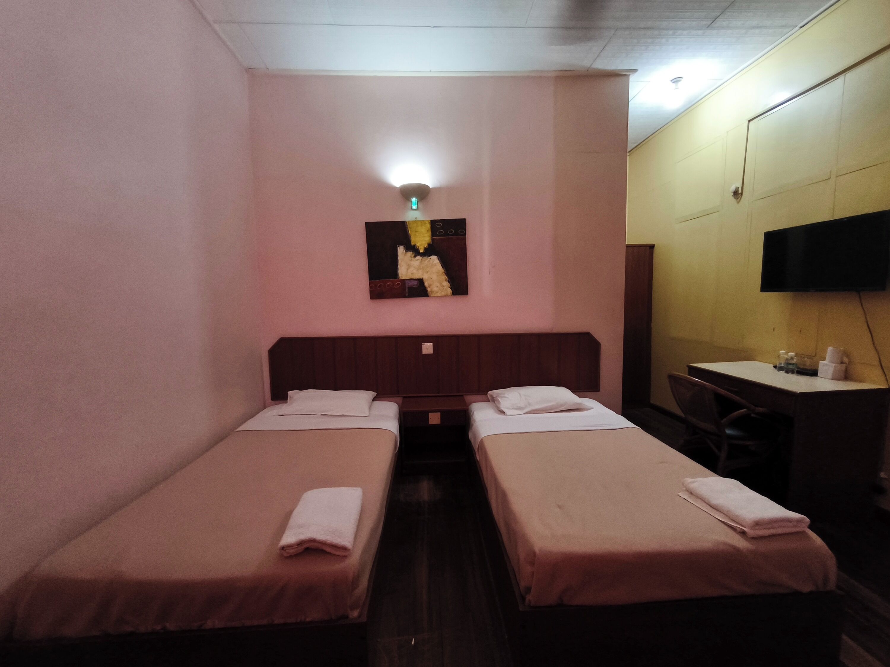 Ein Hotelzimmer mit zwei Einzelbetten, einem Schreibtisch, einem Stuhl, einem an der Wand befestigten Flachbildfernseher und einer Deckenleuchte.