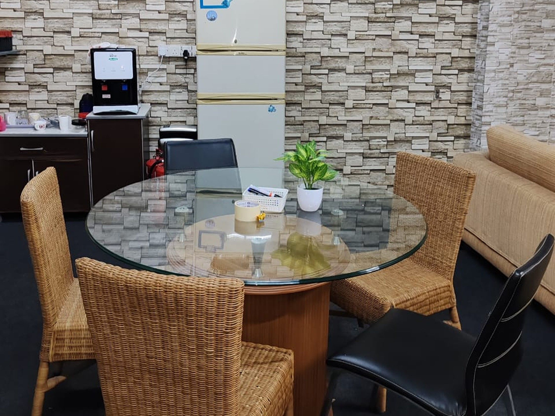 A round glass table with wicker chairs, a potted plant, and a small basket with pens.