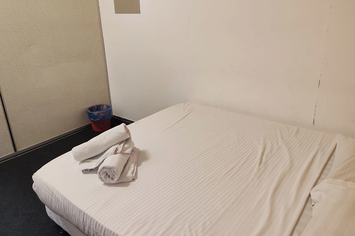 Ein ordentlich bezogenes Bett mit weißen Leinen, ein blauer Mülleimer und ein kleines Fenster in der Wand.