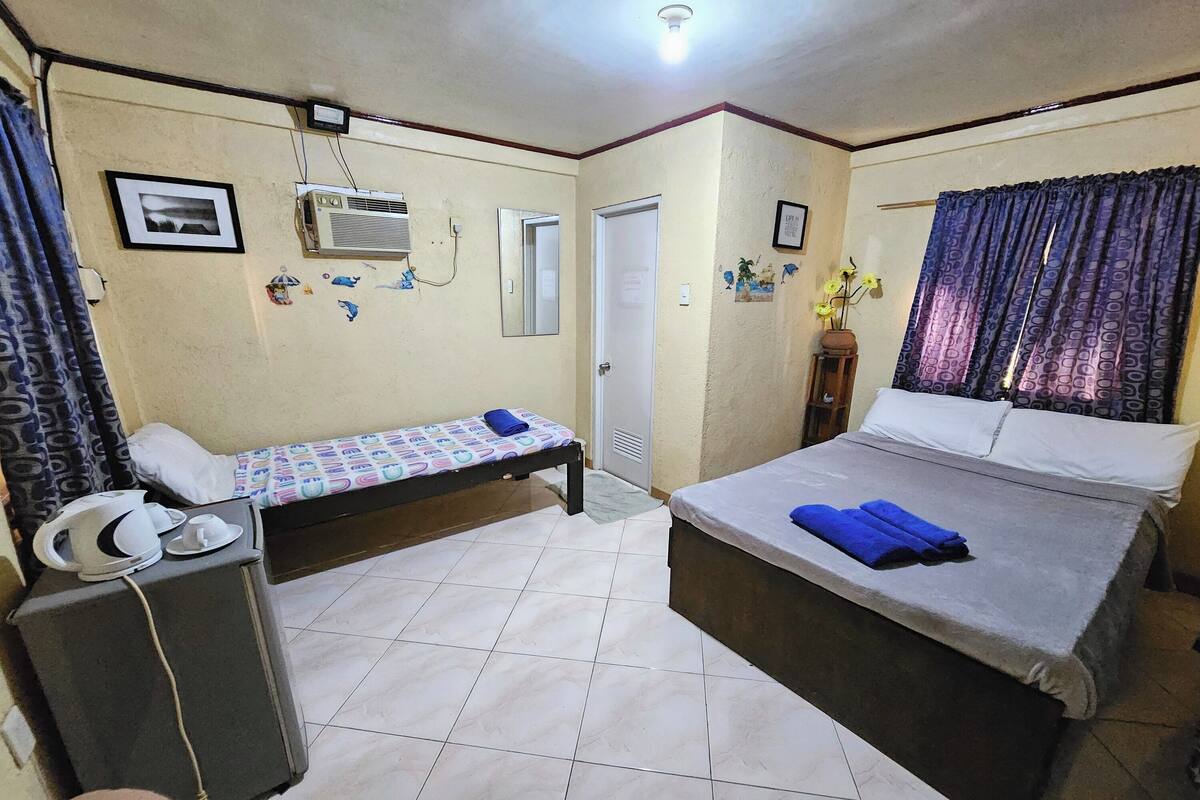 A bedroom with a bed, a bedside table, a small desk with a chair, a heater, and a window with curtains.