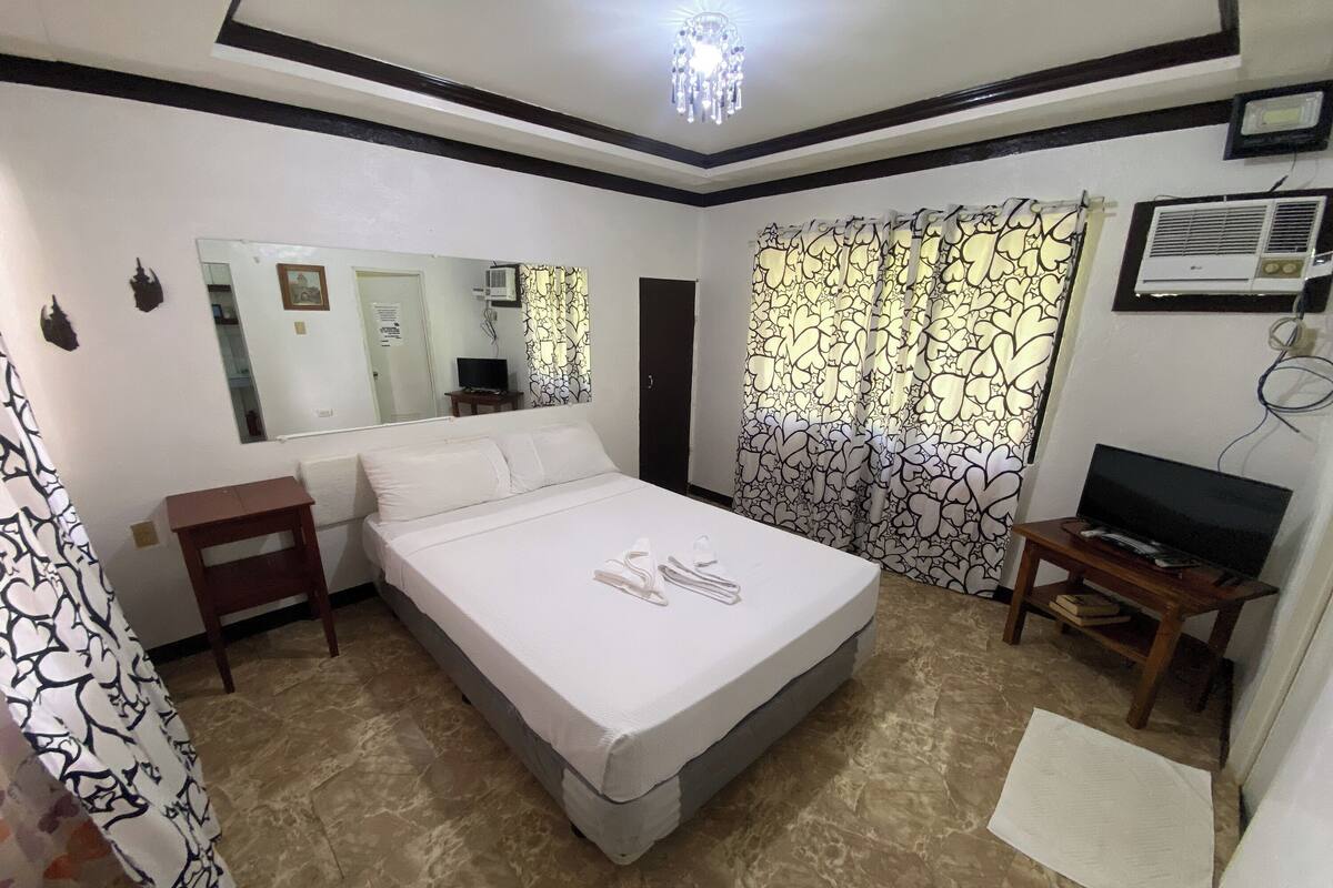 A hotel room with a bed, a TV on a stand, a mirror, a small table, and a curtain with a floral pattern.