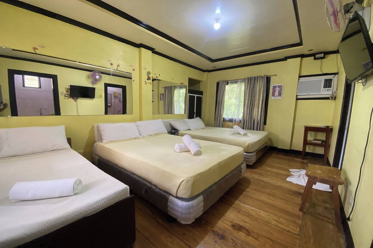 A hotel room with two beds, wooden flooring, a TV, an air conditioning unit, and a small wooden table.