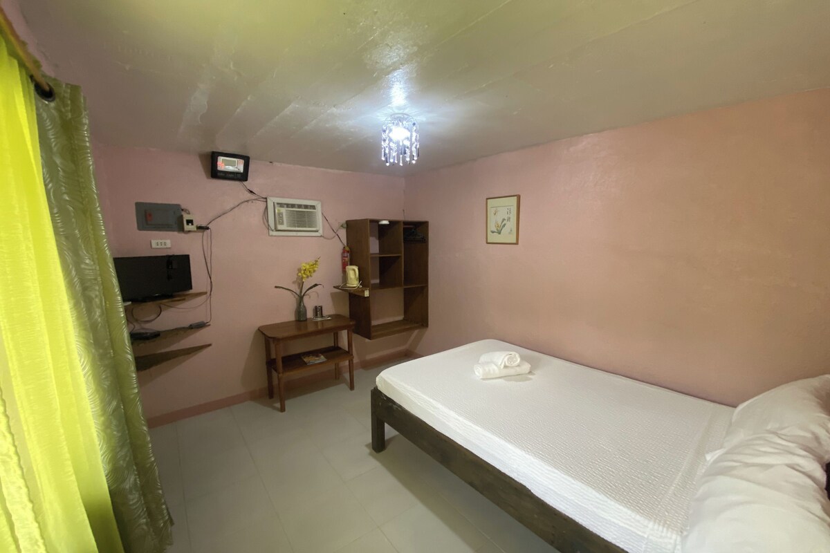 A small, pink-walled room with a bed, a TV, a wooden cabinet, and a small table with a vase.