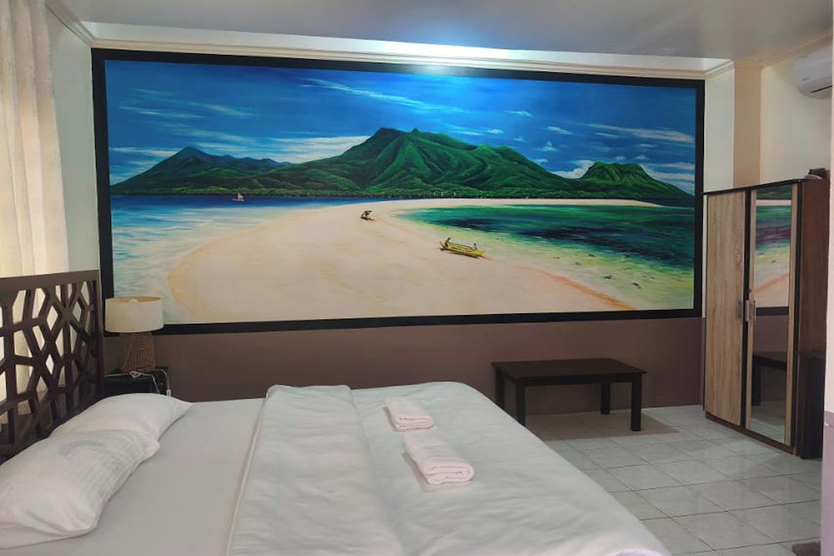 A hotel room with a large beach mural on the wall, a bed with white linens, a small bedside table, and a wooden wardrobe.