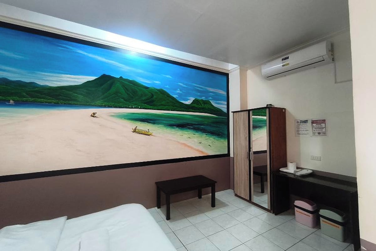 Habitación de hotel con un mural grande de playa, una cama, una mesita pequeña, un escritorio con una computadora y un equipo de aire acondicionado.