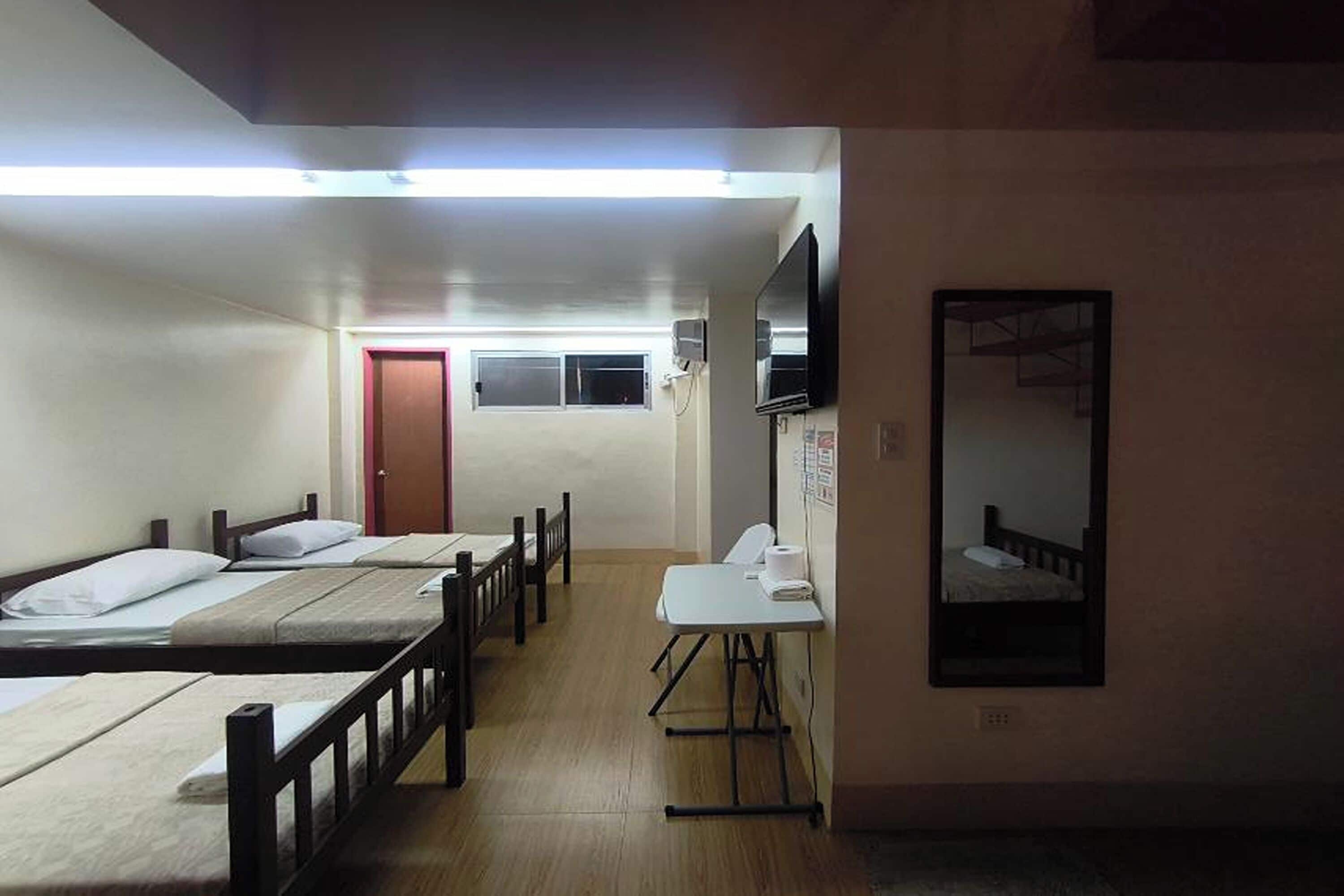 Un dormitorio con cuatro camas individuales, una mesita pequeña, un espejo y una puerta al fondo.