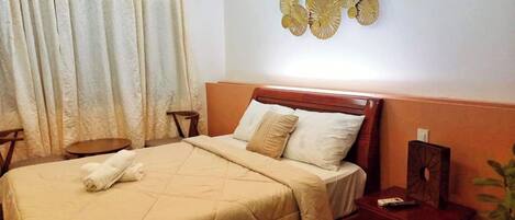 Una cama bien hecha con cubrecama y almohadas color beige, una mesita de noche de madera con un control remoto y un adorno en la pared.