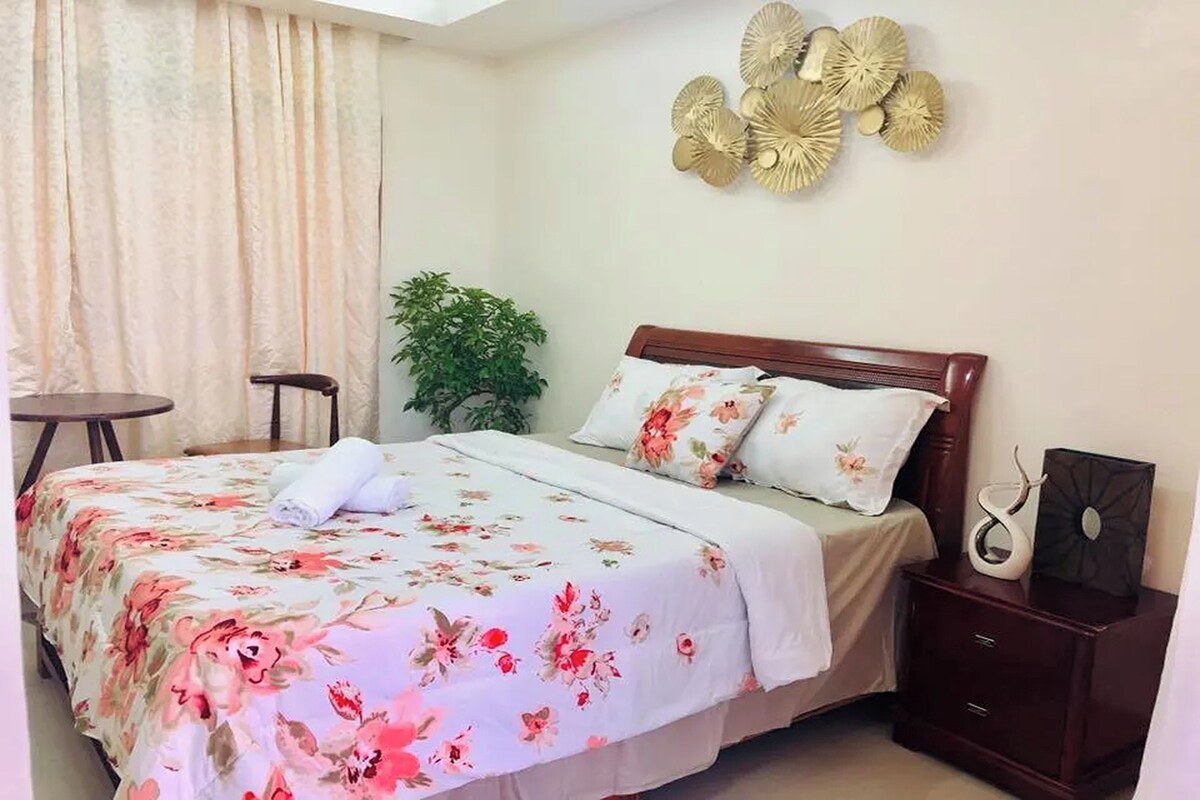 Een opgemaakt bed met bloemenbeddengoed, een nachtkastje met decoratie en een kunstwerk aan de muur.