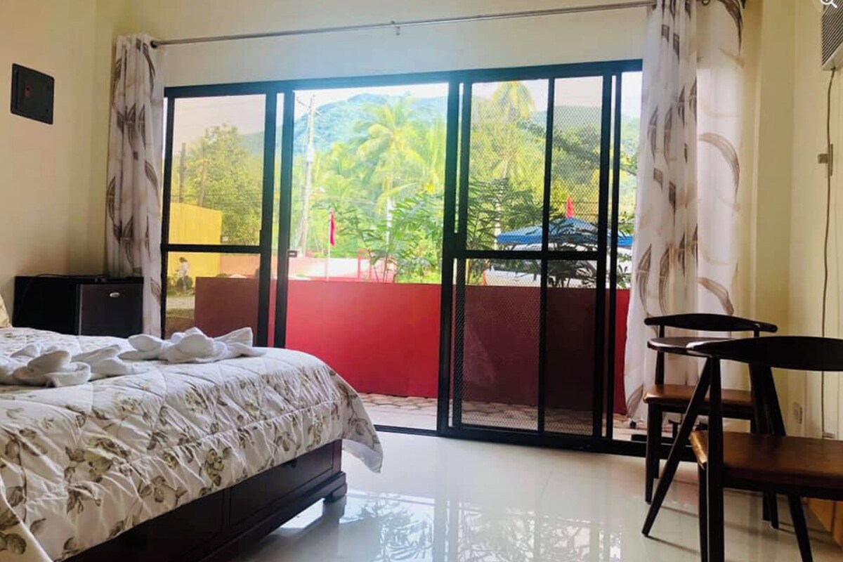 Un dormitorio con cama, escritorio y vistas a un espacio exterior con un muro rojo y vegetación a través de una puerta corrediza de vidrio.