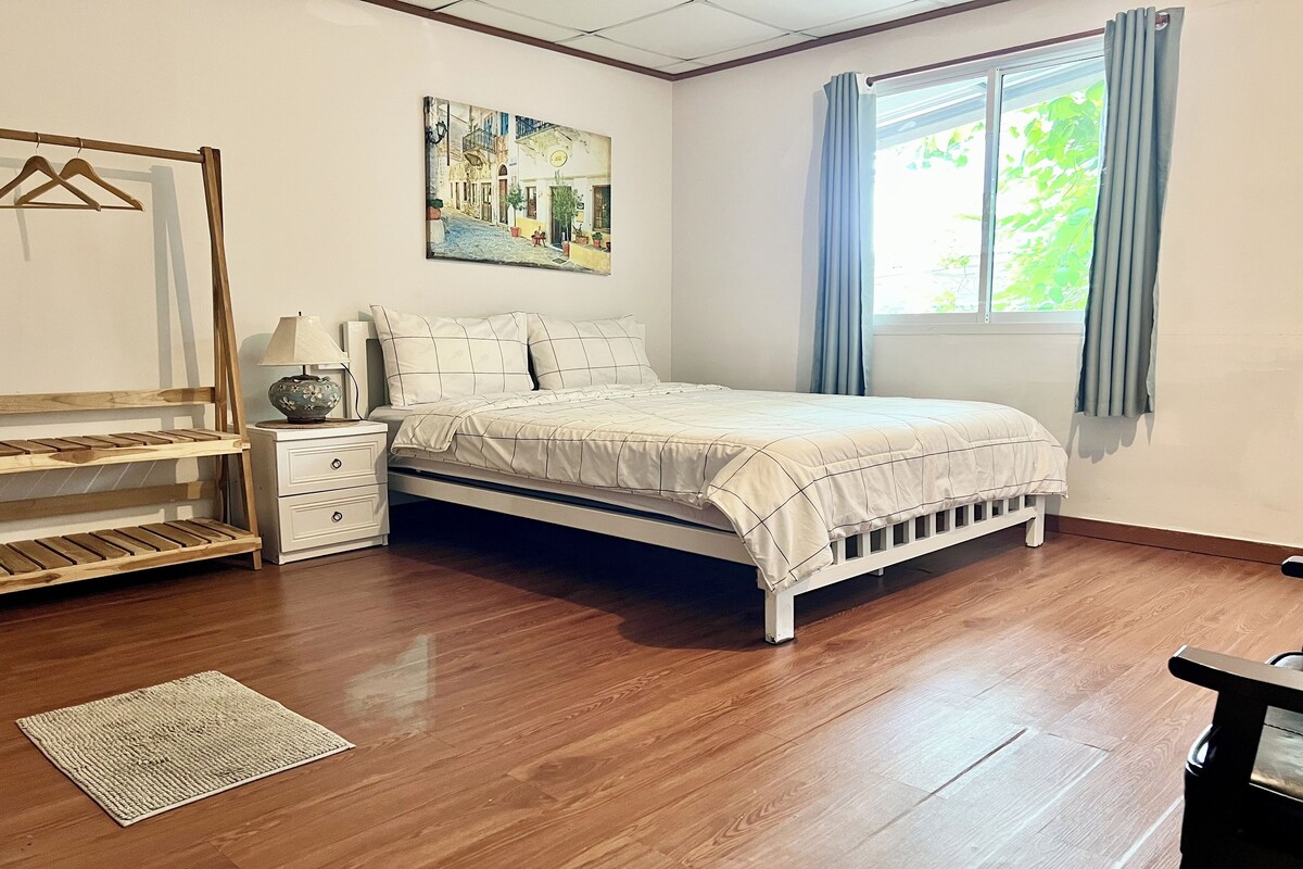 A bedroom with a bed, a nightstand, a lamp, a hanging rack, a painting, and a window with curtains.