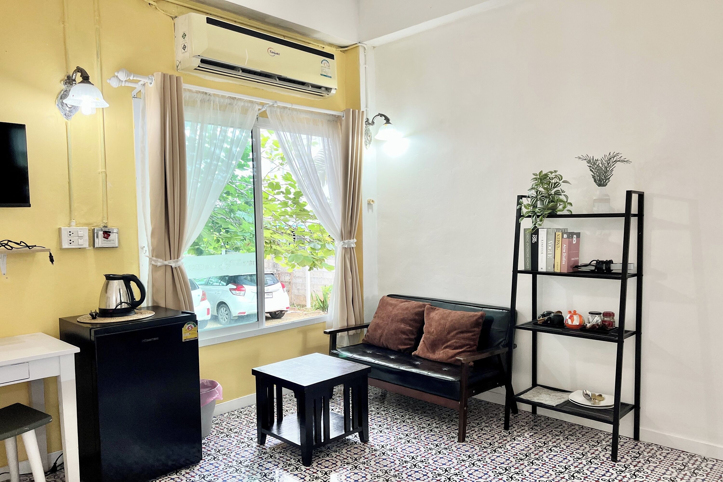 A compact living space with a sofa, a small table, a bookshelf, an air conditioning unit, and a window with curtains.