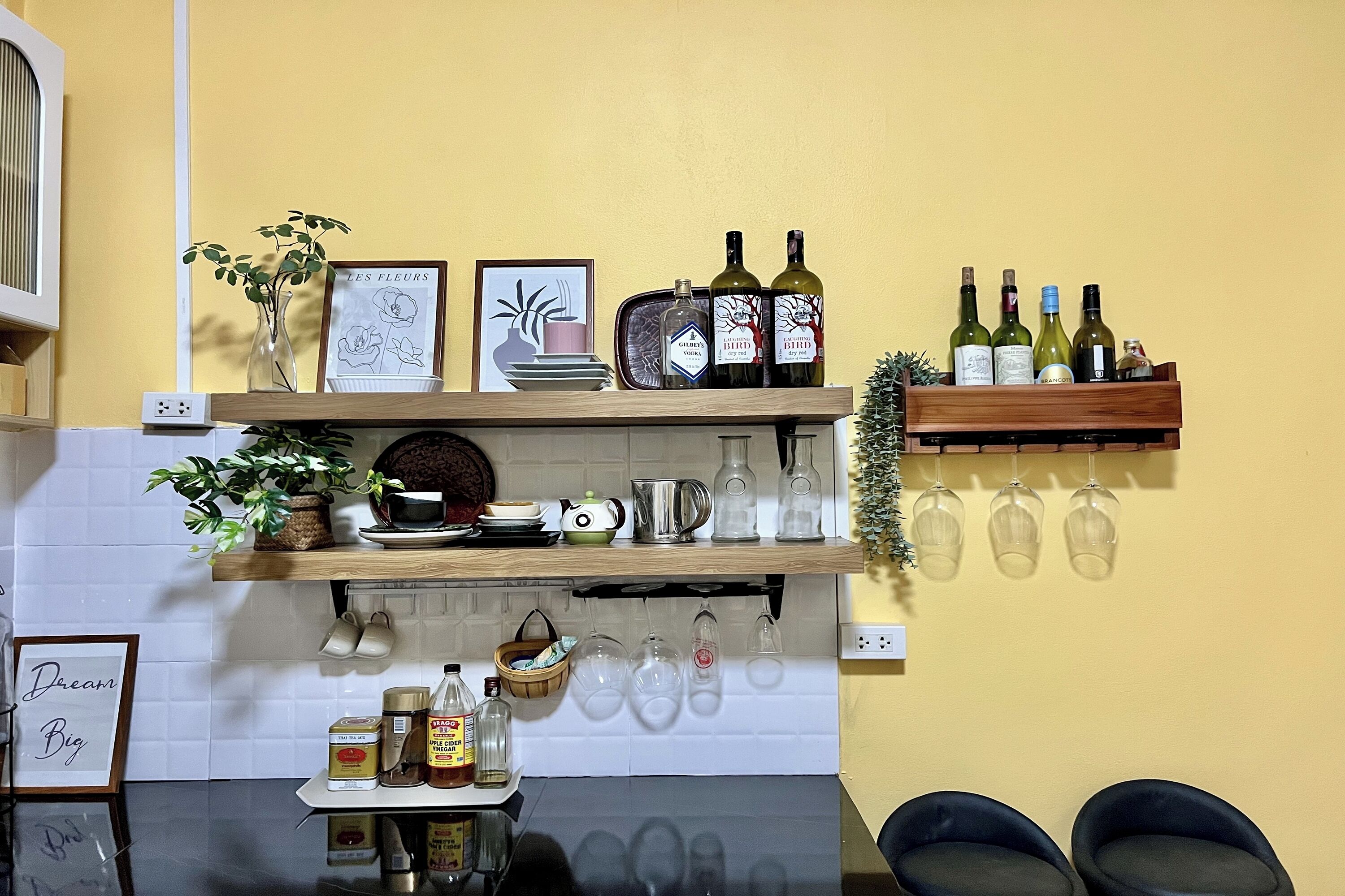 Une cuisine avec des étagères en bois où sont rangés divers objets, notamment des bouteilles, un vase avec des feuilles vertes et des photos encadrées.