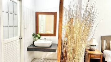 Een badkamer met een wit wastafelmeubel, een spiegel erboven en een houten nachtkastje met een lamp.
