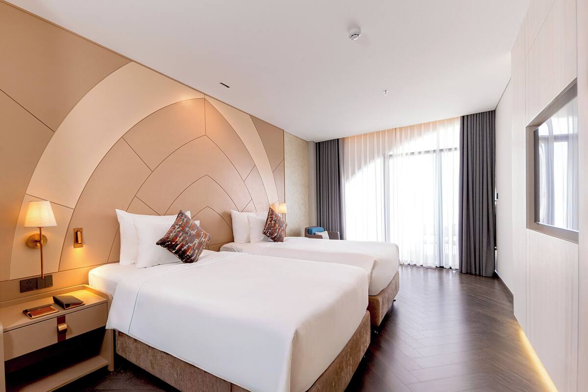 A hotel room with two beds, a nightstand with a lamp, a large arched ceiling, and a view of the outdoors through the window.