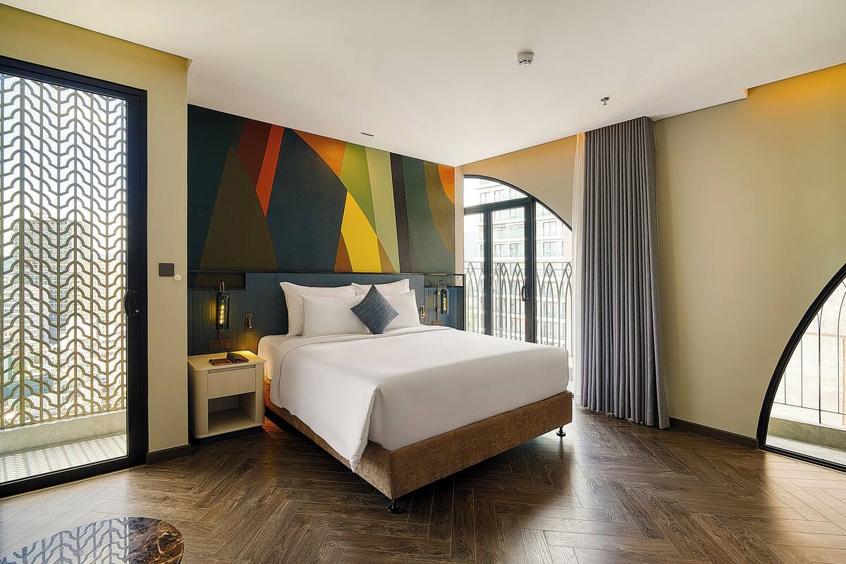 A modern hotel room with a large bed, a nightstand, a decorative wall, and a view of the outside.