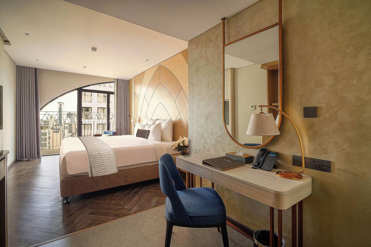 A modern hotel room with a large bed, a desk with a lamp, and a view of the city through a large window.