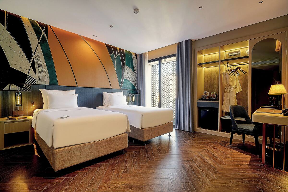 A hotel room with two beds, a desk with a chair, a mirror, and a wardrobe.