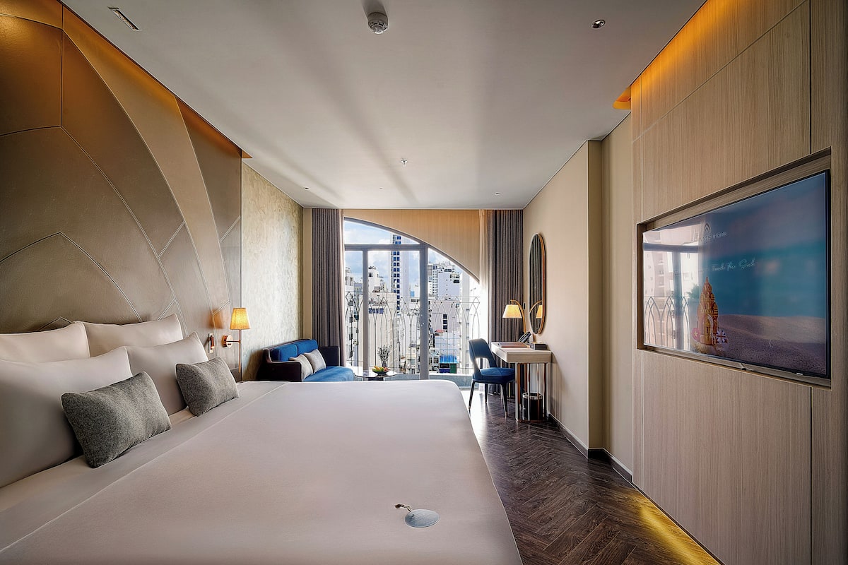 A modern hotel room with a large bed, a desk, a chair, and a large window offering a city view.