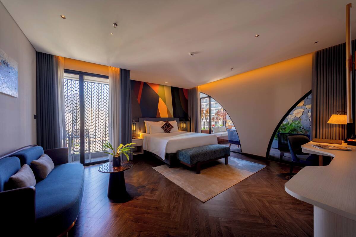 A modern hotel room with a bed, sofa, desk, and a view of the outdoors.