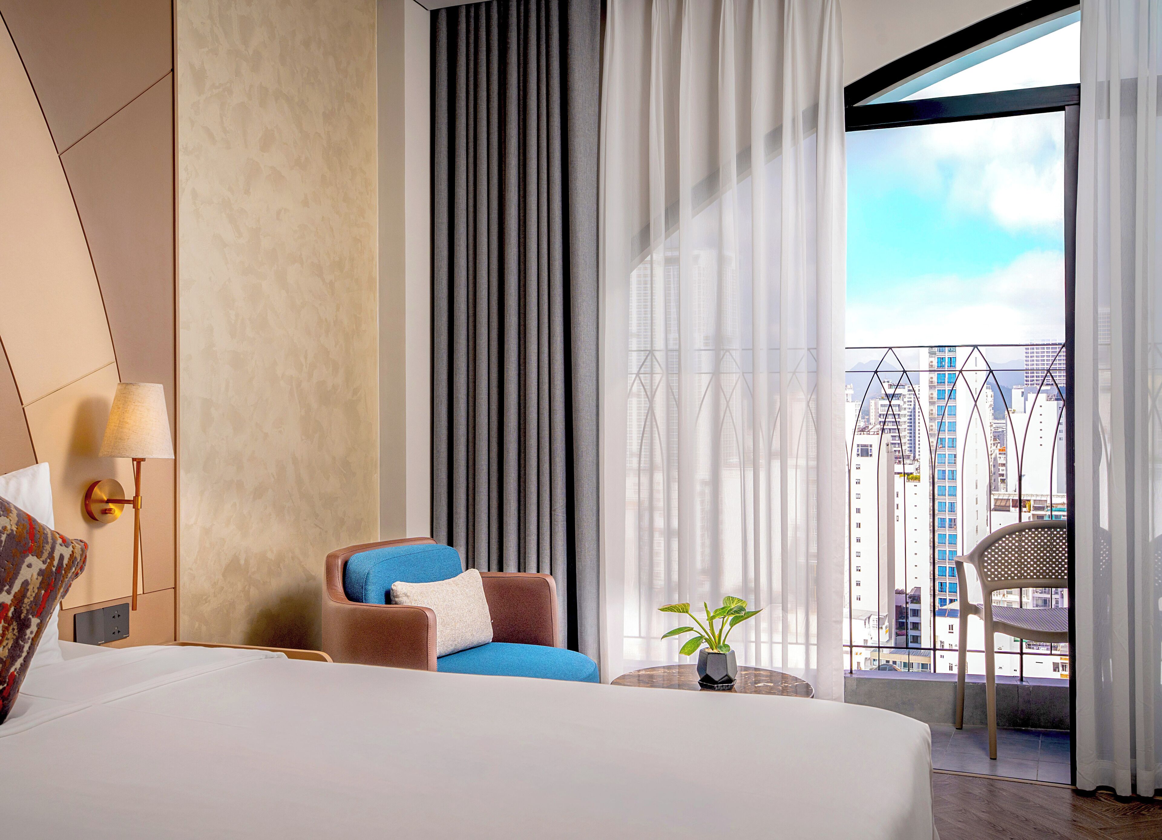 A hotel room with a bed, a chair, a small table with a plant, a lamp, and a view of the city through large windows.
