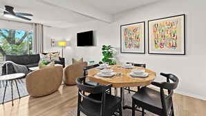 Een moderne woonkamer met een ronde eettafel, zwarte stoelen en een grijze bank. Er is een flatscreen-televisie aan de muur bevestigd en een plafondventilator.