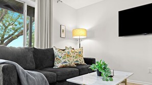 Een moderne woonkamer met een grijze bank, gestreepte kussens, een witte salontafel, een vloerlamp en een potplant.