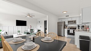 Une cuisine moderne avec des meubles blancs, un four à micro-ondes et un réfrigérateur. On y trouve une table à manger avec de l’assiette et un ventilateur de plafond.