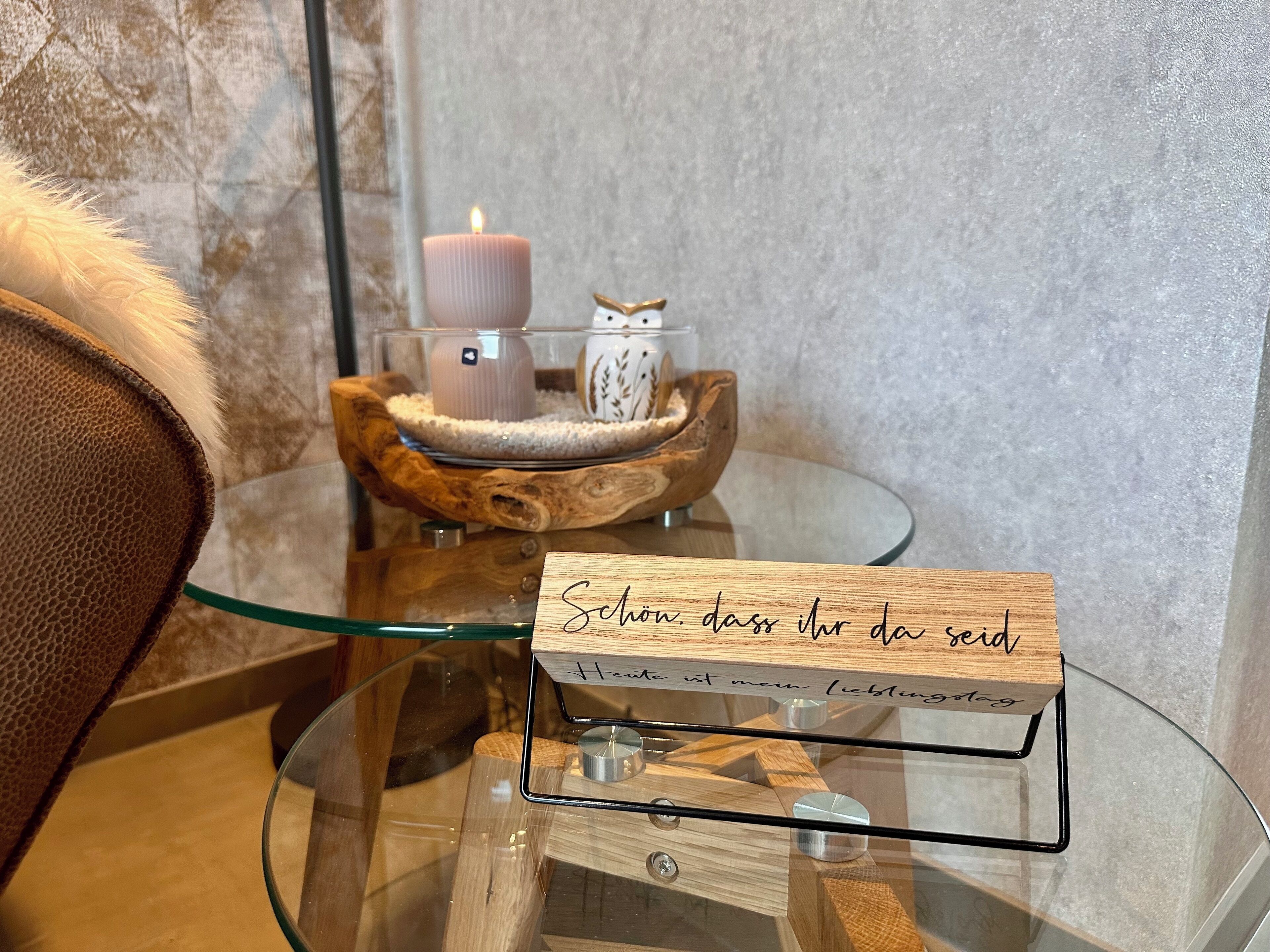 Ein Glastisch mit einer Holzplakette, einer Kerze und einer Eule als Dekoration.