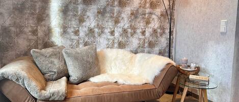 A modern living room with a brown sofa, fluffy cushions, a glass coffee table, and a textured wall.