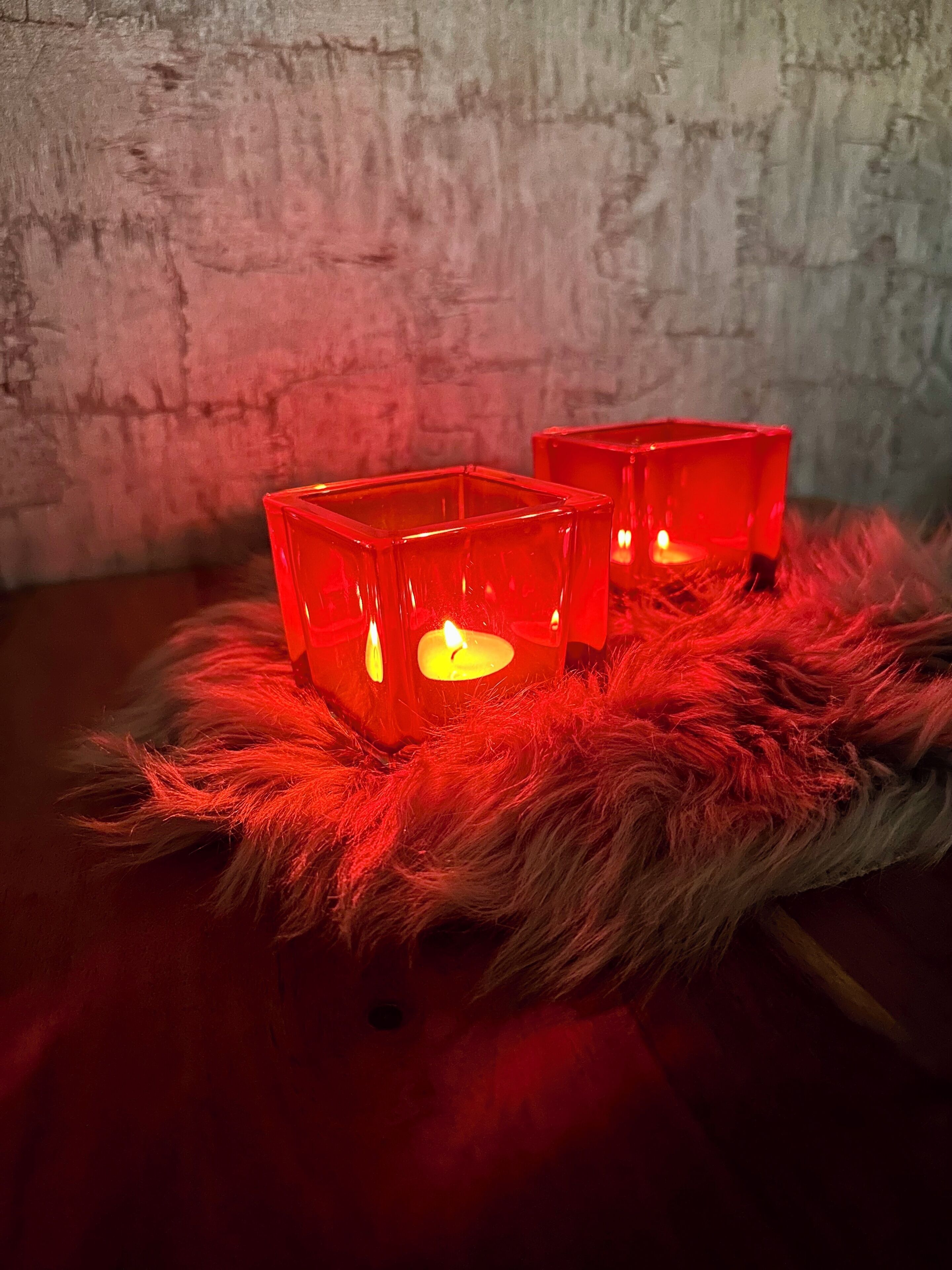 Two red square candle holders with lit candles on a fluffy red surface against a textured wall.