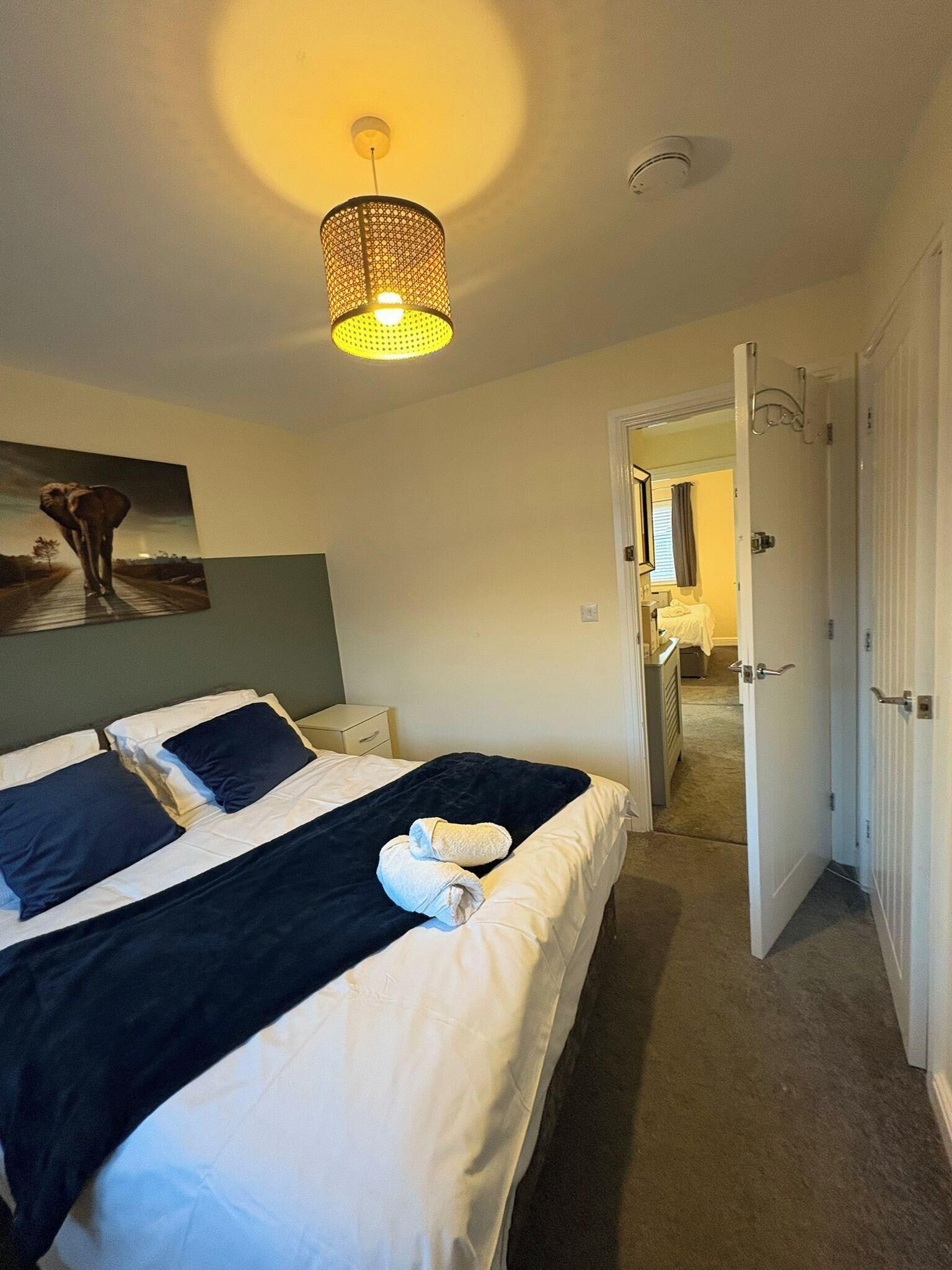 A hotel room with a bed, a painting, a ceiling light, and a door leading to another room.
