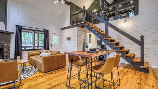 A spacious living room with a wooden floor, a fireplace, a sofa, a coffee table, and a dining area with bar stools. A staircase with a wooden railing leads to an upper level. - Welcome to Tangled Up Cabin! A Luxury Adults Only HotTub/Sauna Playroom (Blue Ridge)
