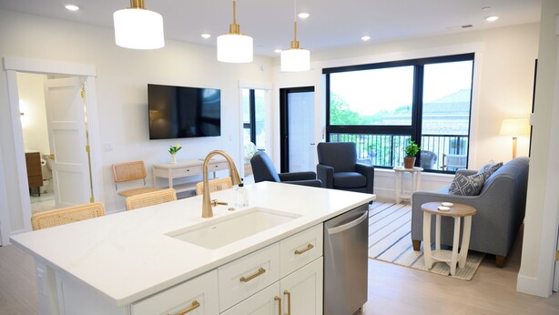Een moderne keuken met een centraal eiland, witte kasten en gouden handgrepen. Er is een geïntegreerde vaatwasser en een groot raam met uitzicht op de omgeving.