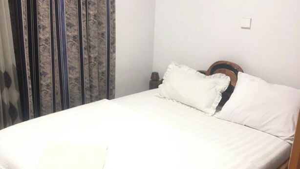 A neatly made bed with white bedding and pillows, a wooden headboard, and a curtain with a leaf pattern. - Lodge- Sleeps 20 - Parking (Kigali)