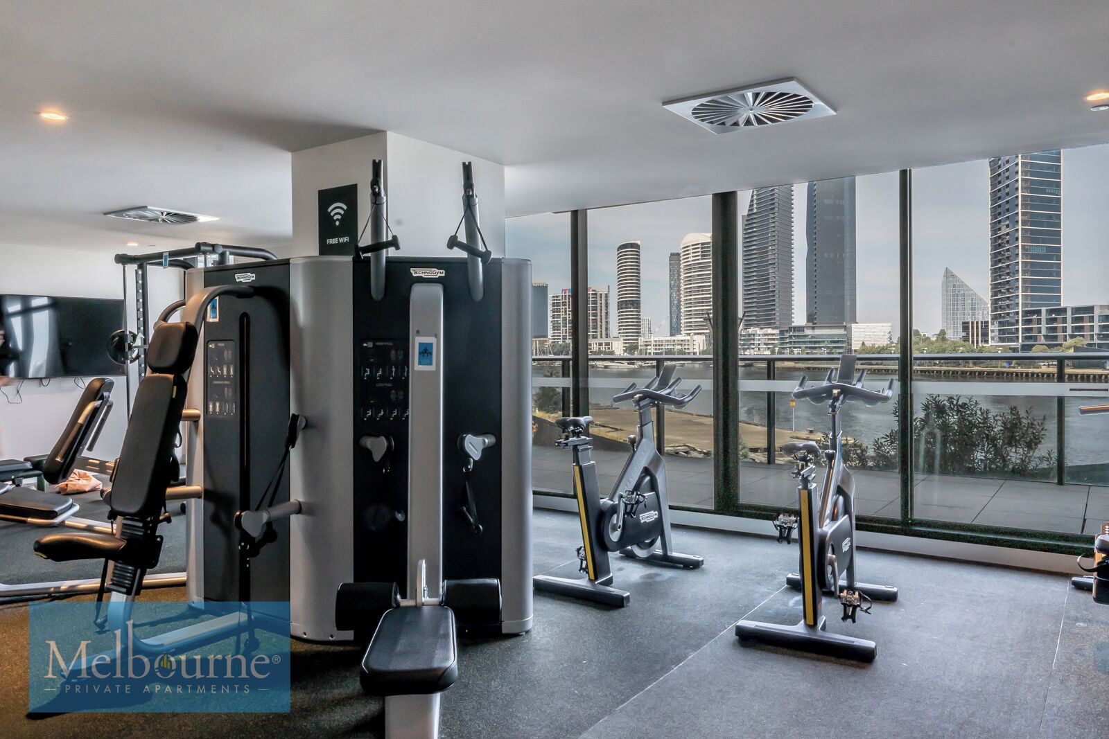 A gym with various exercise equipment, a large window with a city view, and a sign indicating free WiFi.