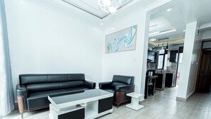 A modern living room with a black leather sofa, a white coffee table, and a small side table. There is a kitchen area in the background with a dining table and chairs.
