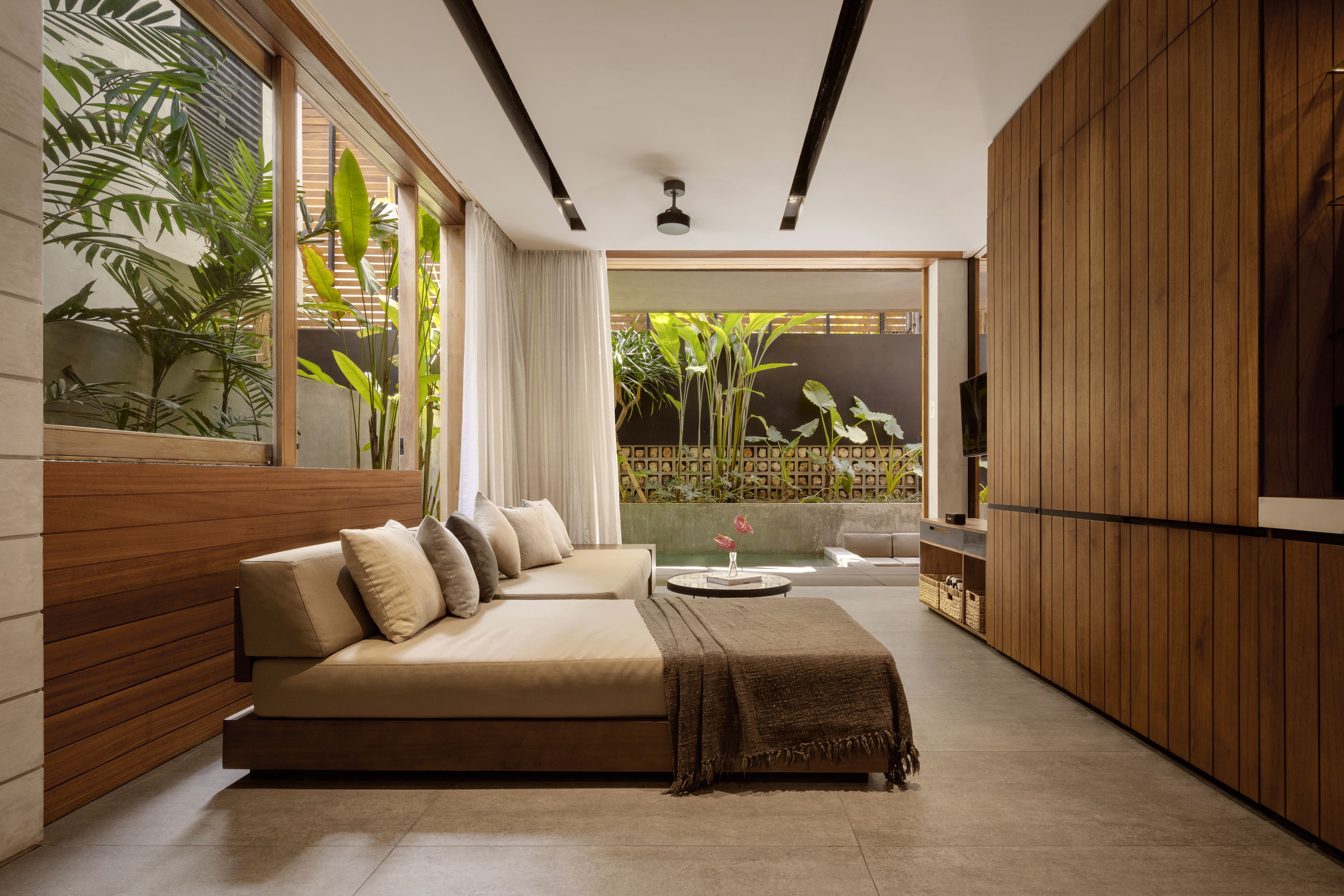 A modern living room with a sofa, a coffee table, and a TV. There are plants and a view of the outdoors.