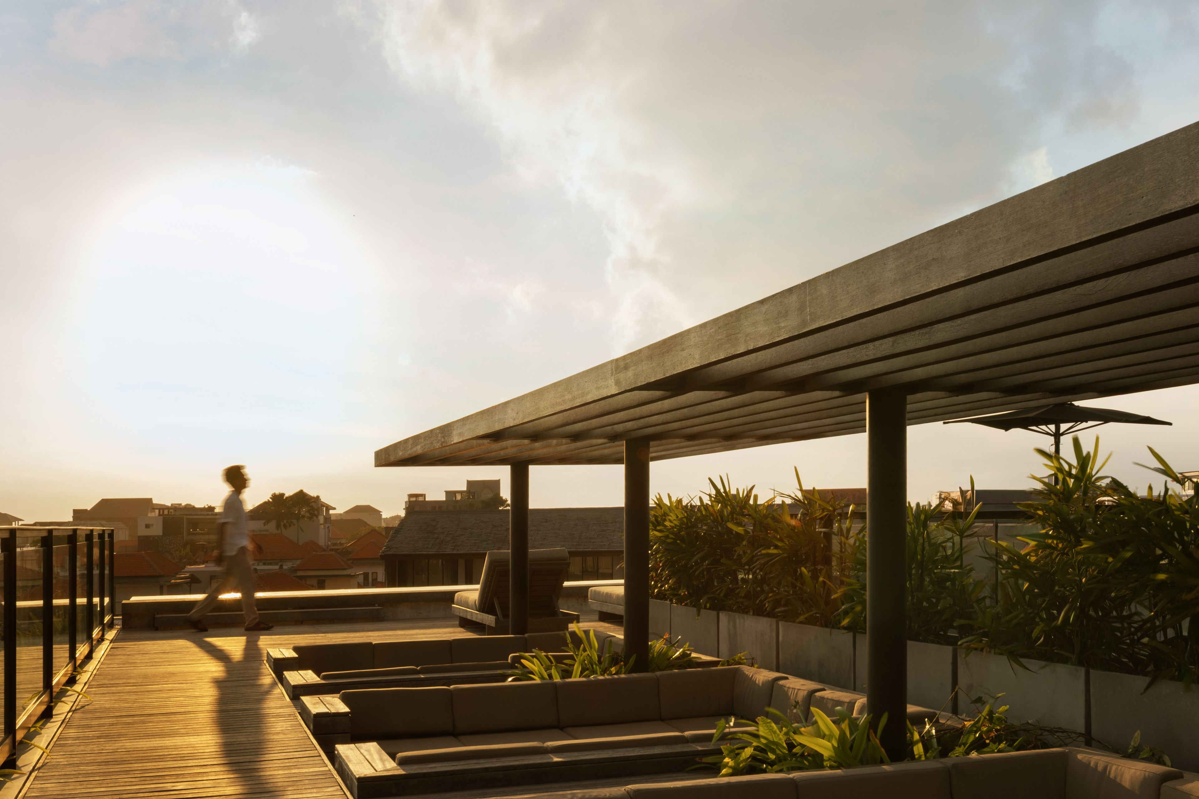 Un área de estar al aire libre con terraza de madera y bancos acolchados, rodeada de follaje y un cielo despejado.