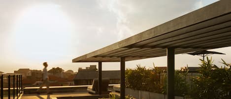 Een buitenruimte met een houten terras en comfortabele banken, omgeven door groen en een heldere lucht.
