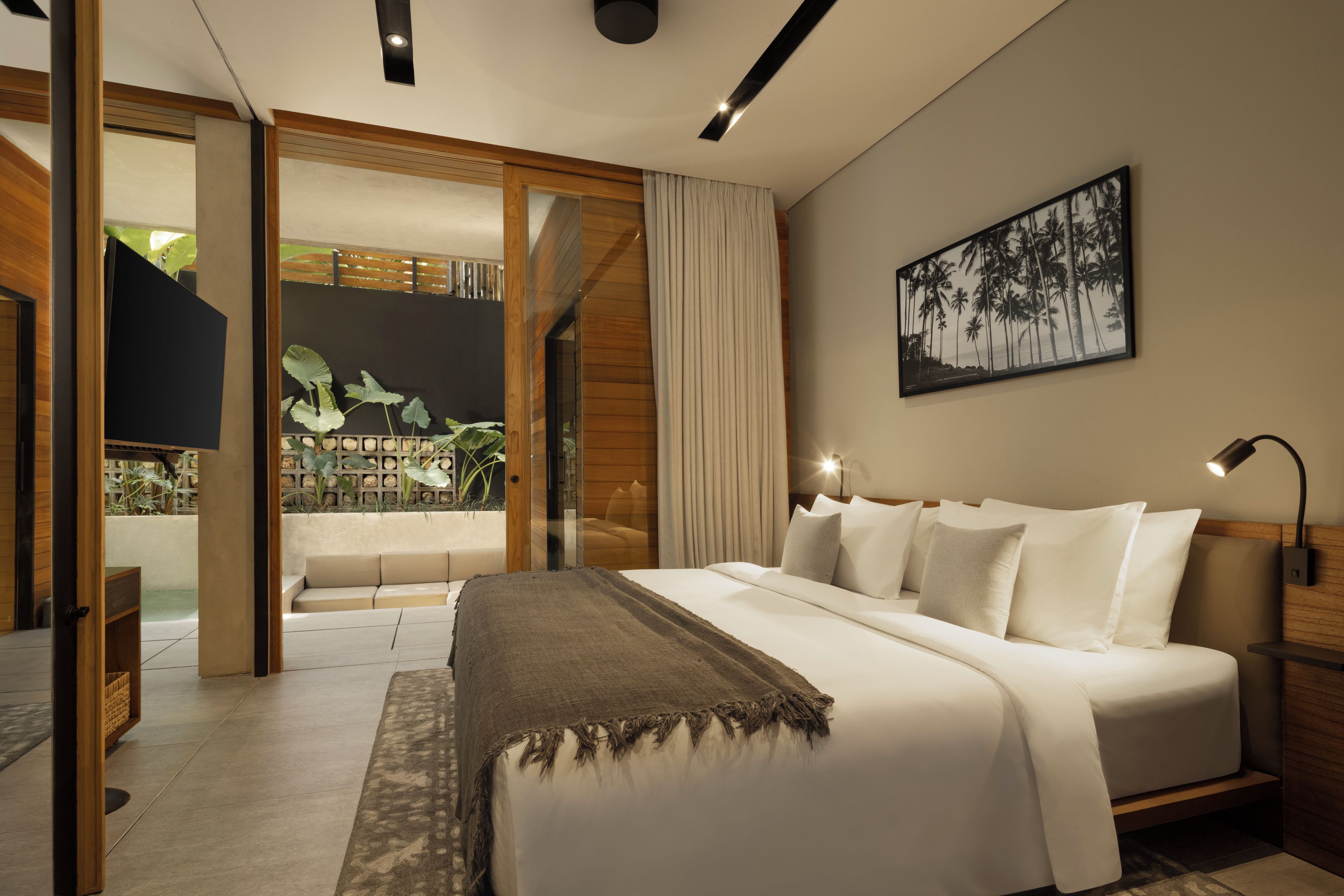 A modern bedroom with a large bed, a nightstand, a wall-mounted lamp, and a view of a balcony with plants.