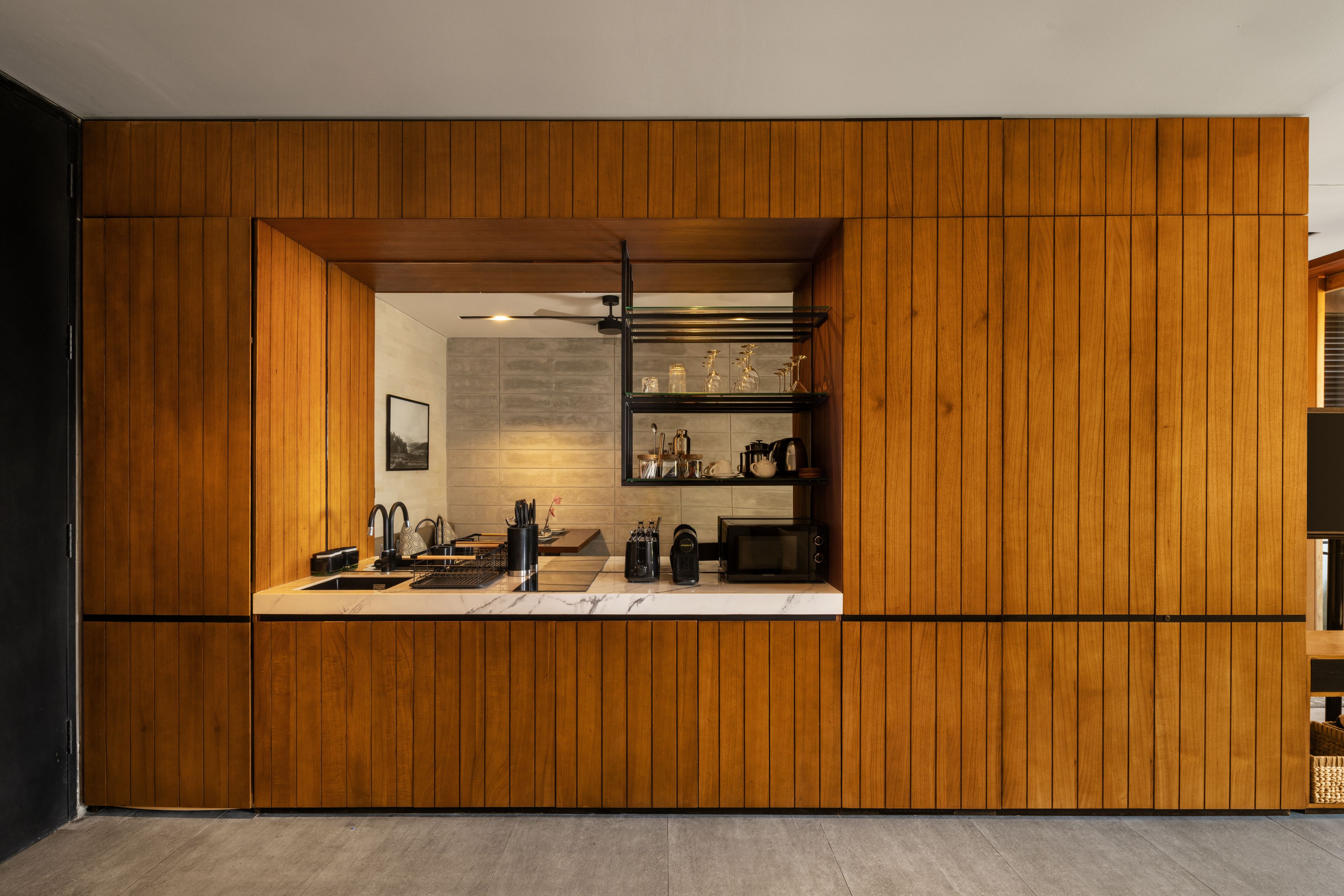A modern kitchen with wooden paneling, a built-in microwave, and a glass shelf with kitchenware.