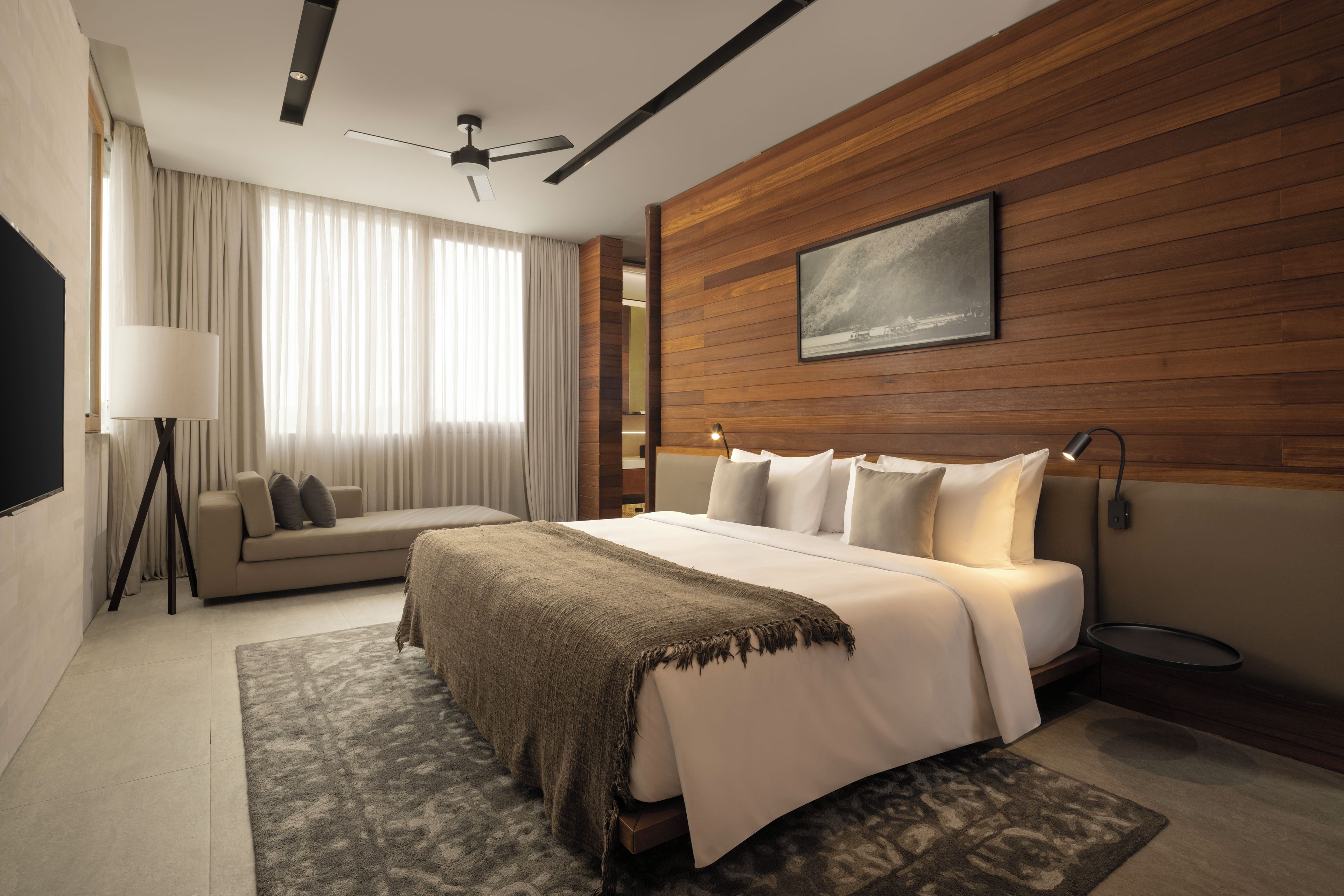 A modern hotel room with a large bed, a wooden accent wall, a ceiling fan, and a large window with curtains.
